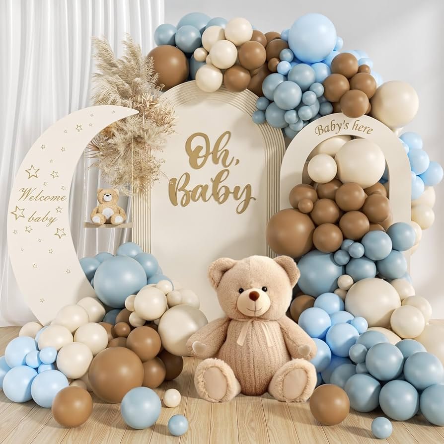A cute baby shower decoration featuring a large teddy bear surrounded by brown, beige, and blue balloons, with crescent moon and arch signs reading “Welcome baby,” “Oh, baby,” and “Baby’s here,” set against a soft, neutral background.